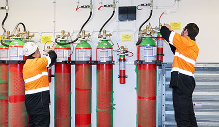 Two men in an orange safety vests stand beside a large group of fire extinguishers during a fire suppression system installation